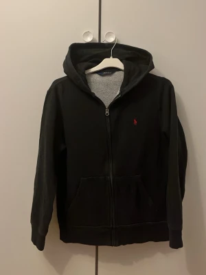 Svart hoodie från Polo Ralph Lauren - Svart hoodie med dragkedja från Polo Ralph Lauren. Klassisk design med huva, två fickor framtill och röd broderad logga på bröstet. Insidan är mjuk och gråmelerad. Perfekt för en chill och stilren look. Passar någon som är runt 160-167