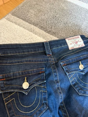 True Religion mörkblå bootcut jeans - Säljer ett par mörkblå jeans från True Religion med bootcut-modell. Jeansen har kontrastsömmar, dekorativa ficklock bak med knappar och klassisk logopatch i midjan. Materialet är denim med snygg tvätt och detaljerade sömmar.