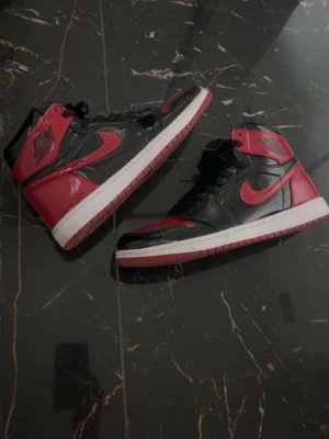 Air Jordan 1 High Bred Patent - Säljer ett par Air Jordan 1 High i den klassiska Bred-färgen med svart och rött patentläder, vit mellansula och röd yttersula. Ikonisk Nike Swoosh på sidorna och Air Jordan-logga vid ankeln. Perfekta för dig som vill sticka ut med en tidlös sneaker. Inga skador eller crease någonstans! Du får i de crease skydd på köpet! Tveka inte på att fråga och priset är inte hugget i en sten!