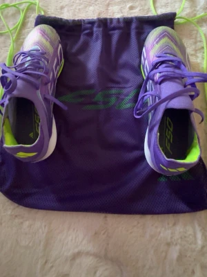 Lila Adidas F50+ Elite fotbollsskor - Säljer ett par lila Adidas F50 fotbollsskor med neongröna detaljer och vita inslag. Skorna har en sportig design och levereras med en matchande lila gympåse med F50-tryck och neongröna snören. Perfekta för dig som vill sticka ut på planen.