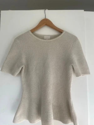 Beige ribbstickad topp med kort ärm - Säljer en beige ribbstickad topp med korta ärmar och rund hals. Tröjan har en lätt utsvängd passform nedtill och är gjord i ett mjukt, tjockare material som känns lyxigt. Perfekt för dig som gillar stilrena och enkla plagg.