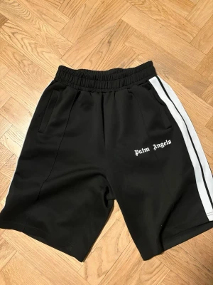 Svarta shorts från Palm Angels - Svarta shorts från Palm Angels med vita sidoränder och logga tryckt på vänster ben. De har elastisk midja och två sidofickor. Perfekta för en avslappnad och sportig stil.