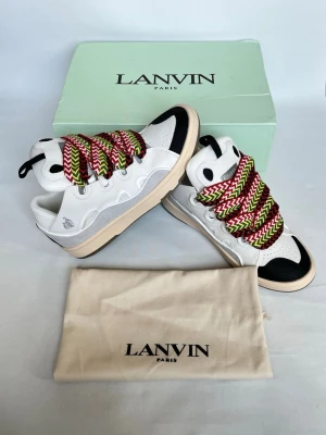 Lanvin vita sneakers med färgglada snören - Lanvin sneakers i vitt läder och mesh med svarta detaljer och chunky sula. Skorna har breda, färgglada skosnören i rött, grönt och vitt zigzag-mönster. Klassisk låg modell med Lanvin-logga på sidan och svart hälflik.