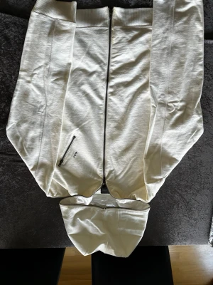 Vit hoodie med dragkedja från IAM - Snygg vit hoodie med hel dragkedja framtill och huva. Har en praktisk ficka med dragkedja och svart IAM-brodyr på sidan. Perfekt för en clean och stilren look. Mjuk bomullskänsla och ribbade muddar vid ärmslut och nederkant.