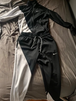 Nike svartvit träningsdress - Snygg svart och vit träningsdress från Nike med dragkedja på jackan och vita detaljer. Setet består av en jacka med hög krage och två fickor samt matchande byxor med dragsko i midjan. Perfekt för träning eller chill. Materialet är mjukt och stretchigt.