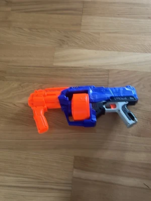 Nerf Elite SurgeFire Blaster - Cool Nerf Elite SurgeFire blaster i blått, orange och grått plastmaterial. Den har en stor roterande trumma för pilar och ett greppvänligt handtag. Perfekt för roliga Nerf-battles med kompisarna.