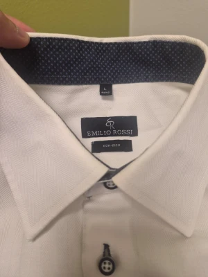 EMILIO ROSSI vit skjorta navyblåa detaljer, storlek L 41/42 - Klassisk vit skjorta från Emilio Rossi i storlek L. Skjortan har lång ärm, knappar framtill och en snygg kontrast på insidan av kragen. Perfekt för en stilren look och enkel att matcha med olika outfits.