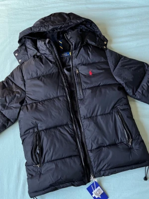 Svart pufferjacka från Polo Ralph Lauren - Snygg svart pufferjacka från Polo Ralph Lauren med huva och dragkedja. Jackan har två stora fickor med dragkedja, en bröstficka och den klassiska röda loggan broderad på bröstet. Perfekt för kalla vinterdagar och riktigt stilren.