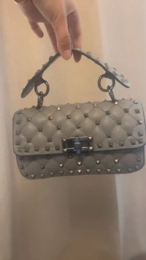 Valentino väska  - Aldrig använd. Snygg grå handväska från Valentino med quiltad design och silverfärgade nitar över hela väskan. Väskan har ett stilrent spänne framtill och ett handtag med nitar för extra edge. Perfekt för dig som gillar att sticka ut med detaljer. Kan ej garanterats. Den har band till. Se r äkta ut. Säljer för 1599