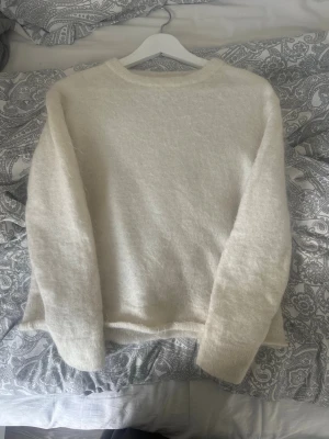 Krämvit fluffig stickad tröja - Mysig krämvit stickad tröja med rund hals och långa ärmar. Tröjan har en fluffig och mjuk struktur som ger en avslappnad och trendig look. Perfekt att matcha med jeans eller kjol för en enkel och stilren outfit.