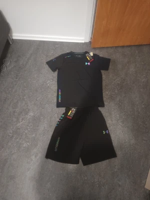 Svart set Under Armour - Snyggt svart set Under Armour med t-shirt och shorts. Båda plaggen har regnbågsfärgat Under Armour-logga och textdetaljer. Tillverkade i ett lätt och funktionellt material som passar perfekt för träning eller chill. Modern och sportig look.
