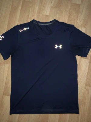 Svart träningströja från Under Armour - Svart kortärmad träningströja från Under Armour med reflekterande logga på bröstet och texten UA-SPEC på axeln. Tillverkad i ett lätt och snabbtorkande material som passar perfekt för intensiva träningspass.