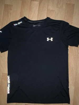 Svart träningströja från Under Armour - Svart kortärmad träningströja från Under Armour med logga på bröstet och text längs sidan. Tillverkad i ett lätt och snabbtorkande material som passar perfekt för intensiva träningspass. Snygg och sportig design med rund hals.