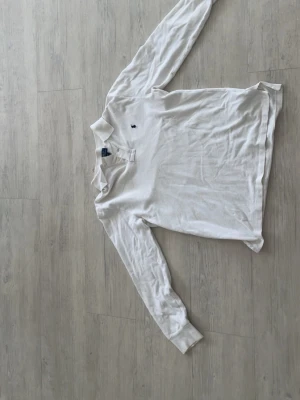 Vit långärmad piké från Polo Ralph Lauren - Klassisk vit långärmad pikétröja från Polo Ralph Lauren med broderad logga på bröstet. Skjortan har krage och knappar vid halsen. Perfekt för en stilren och avslappnad look.