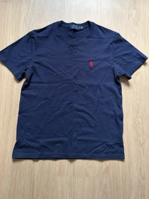 Mörkblå t-shirt från Polo Ralph Lauren - Snygg mörkblå t-shirt från Polo Ralph Lauren med klassisk passform. T-shirten har korta ärmar och är tillverkad i mjuk bomull. På bröstet finns den ikoniska röda Polo-loggan broderad. Perfekt för en stilren och avslappnad look.