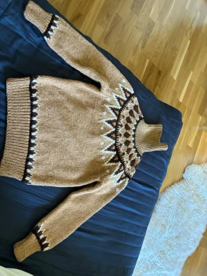 Isländsk tröja Beige stickad tröja med mönster - Stickad tröja i beige med brunt och vitt mönster runt halsen. Tröjan har hög krage, långa ärmar och ribbstickade muddar. Materialet ser ut att vara ull eller ullmix, perfekt för kalla dagar. Klassisk nordisk design som sticker ut.