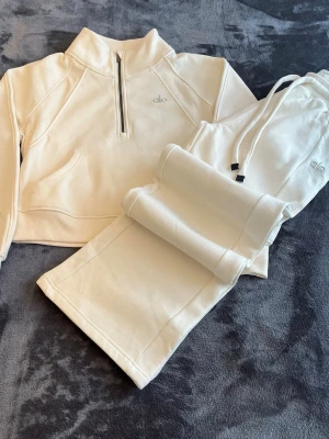 Alo yoga tracksuit - Snygg beige tracksuit från Alo med halv dragkedja framtill och broderad logga på bröstet. Hoodien har en stor magficka och är gjord i mjukt bomullsmaterial. Perfekt för en chill och sportig look.