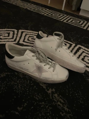 Vita Golden Goose sneakers med stjärna - Snygga vita sneakers från Golden Goose med grå stjärna på sidan och svart detalj vid hälen. Skorna har klassisk låg modell, vita skosnören och är tillverkade i läder. Perfekta för en trendig och avslappnad stil.