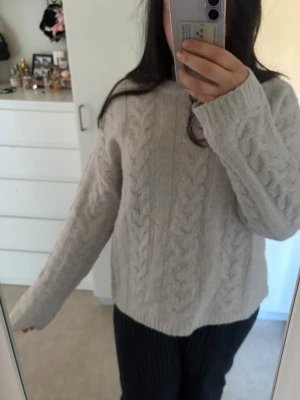 Beige kabelstickad tröja - Mysig beige kabelstickad tröja med rund hals och långa ärmar. Tröjan har ett klassiskt flätmönster och ribbade muddar vid ärmslut och nederkant. Perfekt för lager-på-lager och chill dagar.