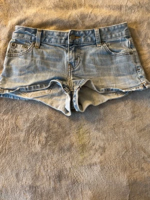Ljusblå jeansshorts från Levi's - Säljer ett par klassiska ljusblå jeansshorts från Levi's med fem fickor, knappgylf och råa benslut. Shortsen har normal passform och är perfekta för varma dagar. Ikonisk Levi's-lapp bak och röd etikett på ena bakfickan.