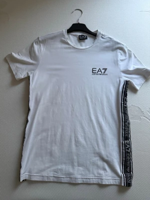 Vit EA7 Emporio Armani t-shirt - Snygg vit t-shirt från EA7 Emporio Armani med svart logga på bröstet och svarta sidopaneler med EA7-tryck längs ena sidan. Klassisk rund hals och korta ärmar. Perfekt för en stilren och sportig look.