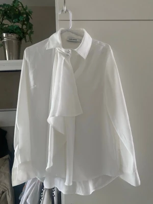 Vit blus med knytband från Carin Wester - Stilren vit blus från Carin Wester med långärmad design och snyggt knytband vid halsen. Klassisk krage och knappar framtill. Perfekt för en trendig och chic look. Materialet känns lätt och luftigt.