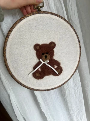 Broderad tavla med brun nalle - Unik nåltovad tavla med en brun nalle i ull,  på ljusbeige bomullstyg och inramad i en rund träbåge. Nallen har en vit rosett runt halsen och detaljer i beige och svart. Perfekt som dekoration till barnrum eller som personlig present.