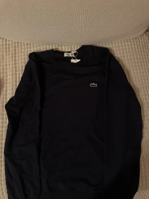 Mörkblå sweatshirt från Lacoste - Snygg mörkblå sweatshirt från Lacoste med klassisk krokodillogga broderad på bröstet. Tröjan har rund halsringning och ribbade muddar vid ärmslut och nederkant. Perfekt för en stilren och avslappnad look.