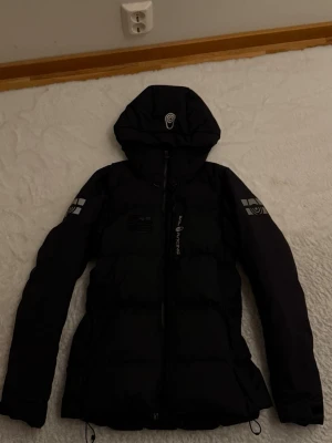 Svart dunjacka från Sail Racing - Snygg svart dunjacka från Sail Racing med huva och dragkedja framtill. Jackan har logga på bröstet och ärmen samt detaljerade tryck. Perfekt för kalla vinterdagar och jackan passar folk som är S o M, de finns bara ett märke och den ser man inte 