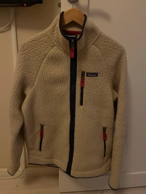 Beige fleecejacka från Patagonia - Säljer en beige fleecejacka från Patagonia med dragkedjor i mörkblått och röda detaljer. Jackan har hög krage, tre fickor med dragkedja och är tillverkad i mjukt teddyfleece-material. Perfekt för kyliga dagar och riktigt skön att ha på sig.