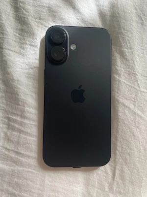 Svart iPhone 16 - Snygg svart iPhone med stilren design och två kameror på baksidan. Telefonen har en matt finish och den klassiska Apple-loggan. Tillverkad i glas och aluminium, perfekt för dig som gillar modern teknik och enkelhet.