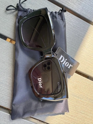 Svarta solglasögon från Dior - Snygga svarta solglasögon från Dior med breda bågar och fyrkantig form. Glasögonen har en glansig finish och tydlig Dior-logga på både glas och skalmar. Perfekt accessoar för att lyfta din stil.