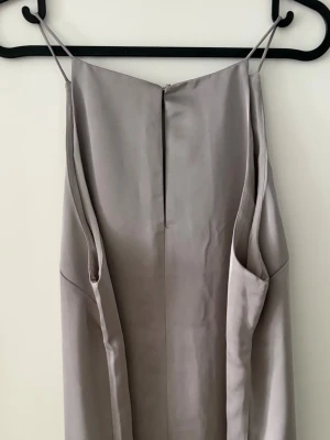 Lång grey ärmlös klänning satin  - Jag har den här satängklänningen från H&M i grått, aldrig använd. Jag har tagit bort etiketten men den är fortfarande helt ny. På bilderna syns några veck efter förvaring, men de försvinner enkelt med ånga/steam. Inga skador alls.
