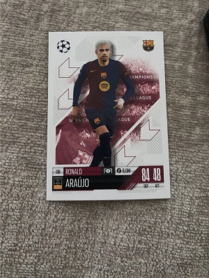 Fotbollssamlarkort Ronald Araújo - Samlarkort med Ronald Araújo från FC Barcelona, UEFA Champions League-serien. Kortet har vit bakgrund med vinröda detaljer och visar spelaren i Barcelonas matchställ. Information om spelarens namn, klubb och statistik finns tryckt på kortet.