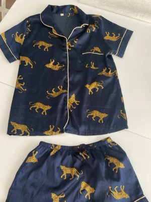 Mörkblå pyjamas med leoparder - Snygg tvådelad pyjamas i mörkblått med kortärmad skjorta och shorts. Setet har ett coolt leopardmönster i gult och vita kontrastkanter. Skjortan har bröstficka och knappar framtill, shortsen har resår i midjan. Perfekt för chillkvällar hemma.