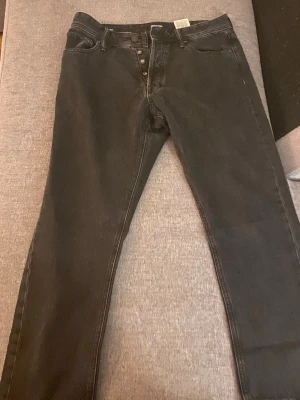 Svarta jeans från J&J - Svarta jeans med rak passform och knappgylf. Klassisk femficksmodell i denim, perfekt för en avslappnad stil. Jeansen har en enkel design utan slitningar eller dekorationer.