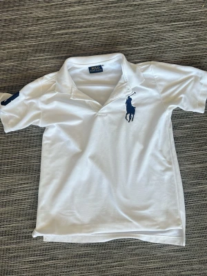 Vit pikétröja från Polo Ralph Lauren - Vit pikétröja från Polo Ralph Lauren med klassisk krage och korta ärmar. Stor marinblå logga på bröstet och siffra på ärmen. Perfekt för en clean och sportig stil.
