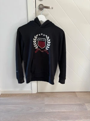 Svart hoodie från GANT med emblem - Snygg svart hoodie från GANT med stort broderat emblem på bröstet. Klassisk känguruficka framtill och huva. Tillverkad i mjuk bomullsblandning som är skön att ha på sig. Perfekt för en avslappnad och sportig stil.