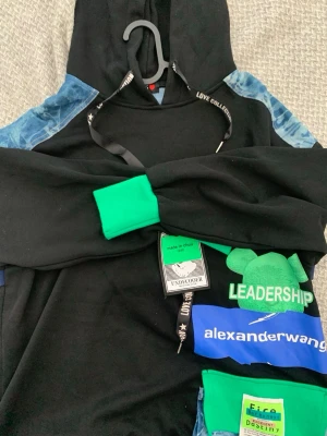 Svart hoodie Alexander Wang x Undercover - Cool svart hoodie med blå och gröna detaljer, tryck med texten 'LEADERSHIP' och 'alexanderwang' samt grönt björnmotiv. Huva med svarta snören med text. Unika patches och etiketter framtill. Perfekt för dig som gillar streetwear och statement-plagg.