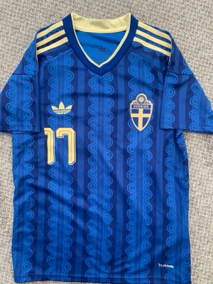 Blå Sverige fotbollströja Adidas - Säljer en blå fotbollströja från Adidas med Sveriges landslagsemblem och nummer 17. Tröjan har gula detaljer, V-ringning och mönstrade ränder. Namnet Gyökeres står på ryggen. Tillverkad i lätt och ventilerande material (Climacool). Perfekt för match eller träning.