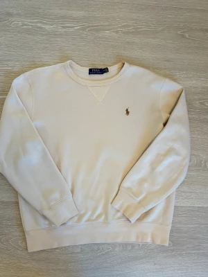 Beige sweatshirt från Polo Ralph Lauren - Snygg beige sweatshirt från Polo Ralph Lauren med klassisk broderad logga på bröstet. Tröjan har rund halsringning, ribbade muddar och är tillverkad i mjukt bomullsmaterial. Perfekt för en avslappnad och stilren look.