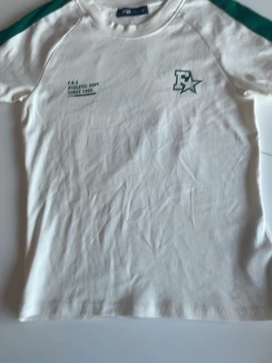 Vit t-shirt från FB Sister med gröna detaljer - Vit t-shirt från FB Sister  New Yorker med gröna axelpartier och tryck på bröstet. Klassisk rund halsringning och korta ärmar. Skön passform och tillverkad i mjuk bomull, perfekt för en avslappnad stil.  Strl M men mer som S 