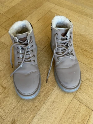 Beige fodrade snörstövlar - Snygga beige stövlar med snörning och mjukt vitt foder. Skorna har en robust sula och är perfekta för kyligare dagar. De har en klassisk design med rund tå och detaljer i brunt vid plösen. Passar dig som vill ha både stil och komfort.