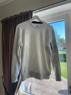  C.P. Company - Vit sweatshirt från C.P. Company med rund halsringning och ribbade muddar. Klassisk modell med en liten ficka och glaslins-detalj på ena ärmen. Perfekt för en clean och stilren look. Sänkt pris på grund av att jag inte har möjlighet att skicka med cp company märket.