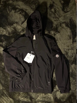 Svart Moncler vindjacka - Svart vindjacka från Moncler med huva och dragkedja framtill. Jackan har två fickor med dragkedja, elastiska muddar och Moncler-logga på ärmen. Tillverkad i lätt och slitstarkt material, perfekt för blåsiga dagar. Skickar samma dag rekommenderar PostNord går oftast snabbast. Skriv om du har frågor svarar så fort jag kan. Nfc tag funkar👍