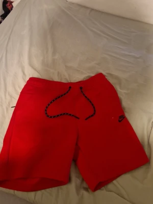 Röda Nike shorts med snörning - Röda shorts från Nike med svartvitt snöre i midjan och klassisk Nike-logga på vänster ben. Perfekta för sommaren och har en avslappnad passform. Tillverkade i mjukt material som känns skönt mot huden.