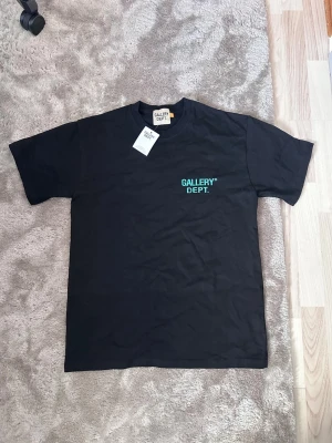 Svart t-shirt från Gallery Dept. - Svart t-shirt från Gallery Dept. med grön logga tryckt på bröstet och stor text på ryggen. Klassisk passform med rund hals och korta ärmar. Perfekt för dig som gillar streetwear och vill sticka ut med en snygg logga.