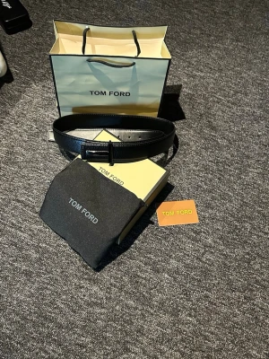 Svart skärp från Tom Ford i skinn - Snyggt svart skärp från Tom Ford i äkta skinn med klassiskt T-spänne i metall. Perfekt accessoar för att lyfta din outfit med en exklusiv touch. Levereras med originalpåse och förpackning.