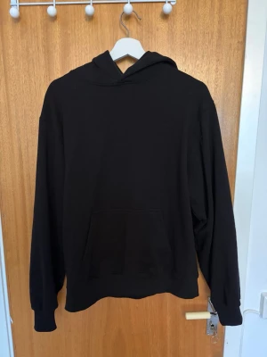 Svart basic hoodie - En stilren svart hoodie med huva och stor magficka framtill. Hoodien har långa ärmar, ribbade muddar och är tillverkad i mjukt material, perfekt för en avslappnad look. Passar dig som gillar enkel och clean streetstyle.