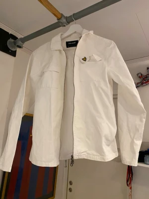 Vit overshirt från Lyle & Scott - Stilren vit overshirt från Lyle & Scott med dragkedja framtill och klassisk krage. Bröstficka med den ikoniska gula fågel-loggan. Perfekt lager på lager-plagg för en clean look. Tillverkad i bomull för en skön känsla.